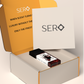 Corner view of a packaging within it a minimalist clear glass perfume bottle with black wood cap containing SERO fragrance inspired by Baccarat Rouge. 